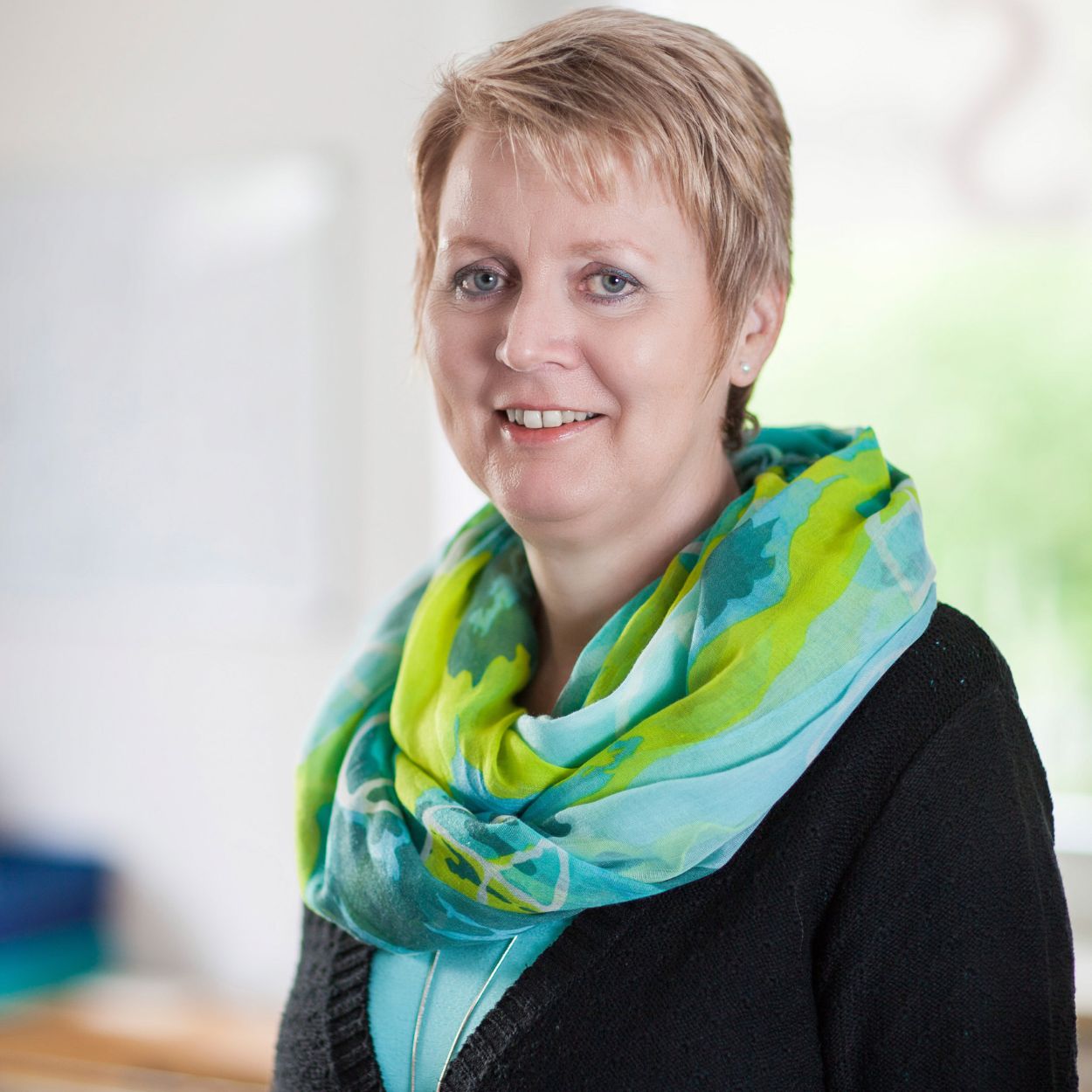 Eine Frau mit kurzen blonden Haaren trägt einen bunten Schal und lächelt in die Kamera.